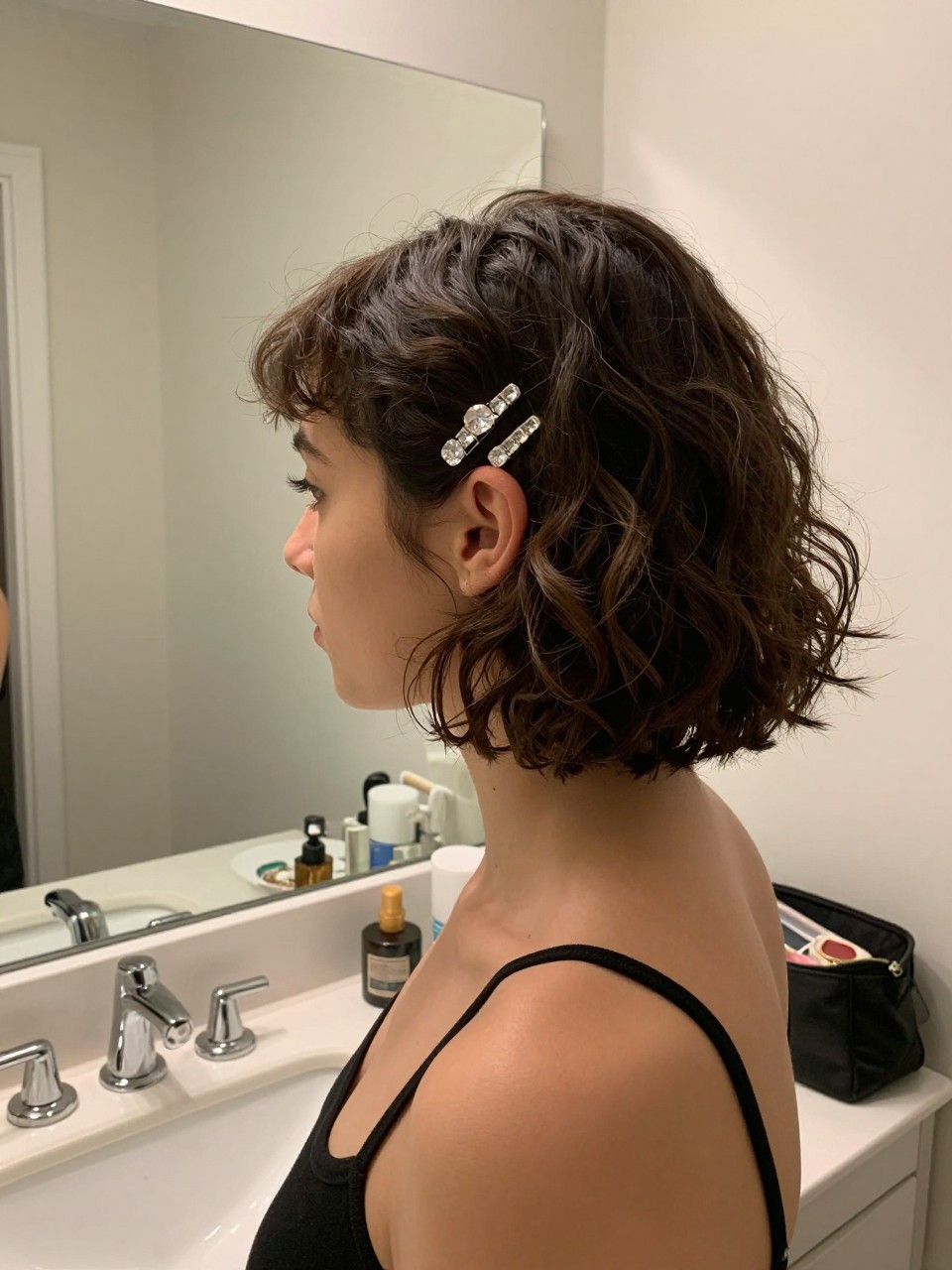 Photo of a woman wearing a softly curled short bob with crystal side pins stacked above one ear, side profile view, setting in a bathroom vanity mirror, lighting soft indoor lighting, containing a small lived-in detail such as a jewelry dish on the counter and a makeup bag nearby, latest iPhone photo quality.