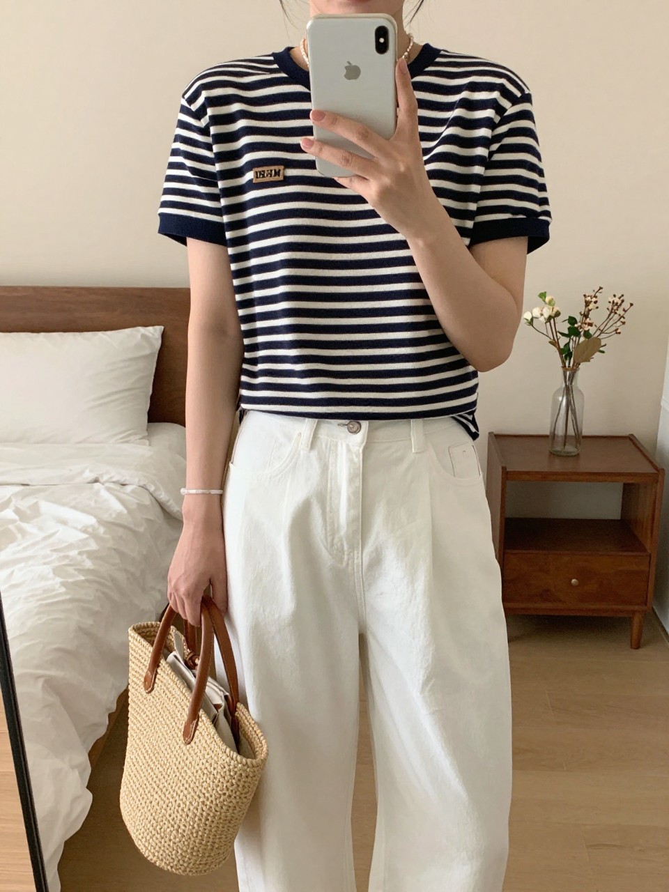 iPhone mirror selfie of a woman in a navy striped Breton top tucked into white wide leg pants holding a straw tote, standing with phone at waist level, cropped at NECK so face doesn't show, in a sunlit feminine-themed bedroom, warm moody lighting, iPhone photo quality.