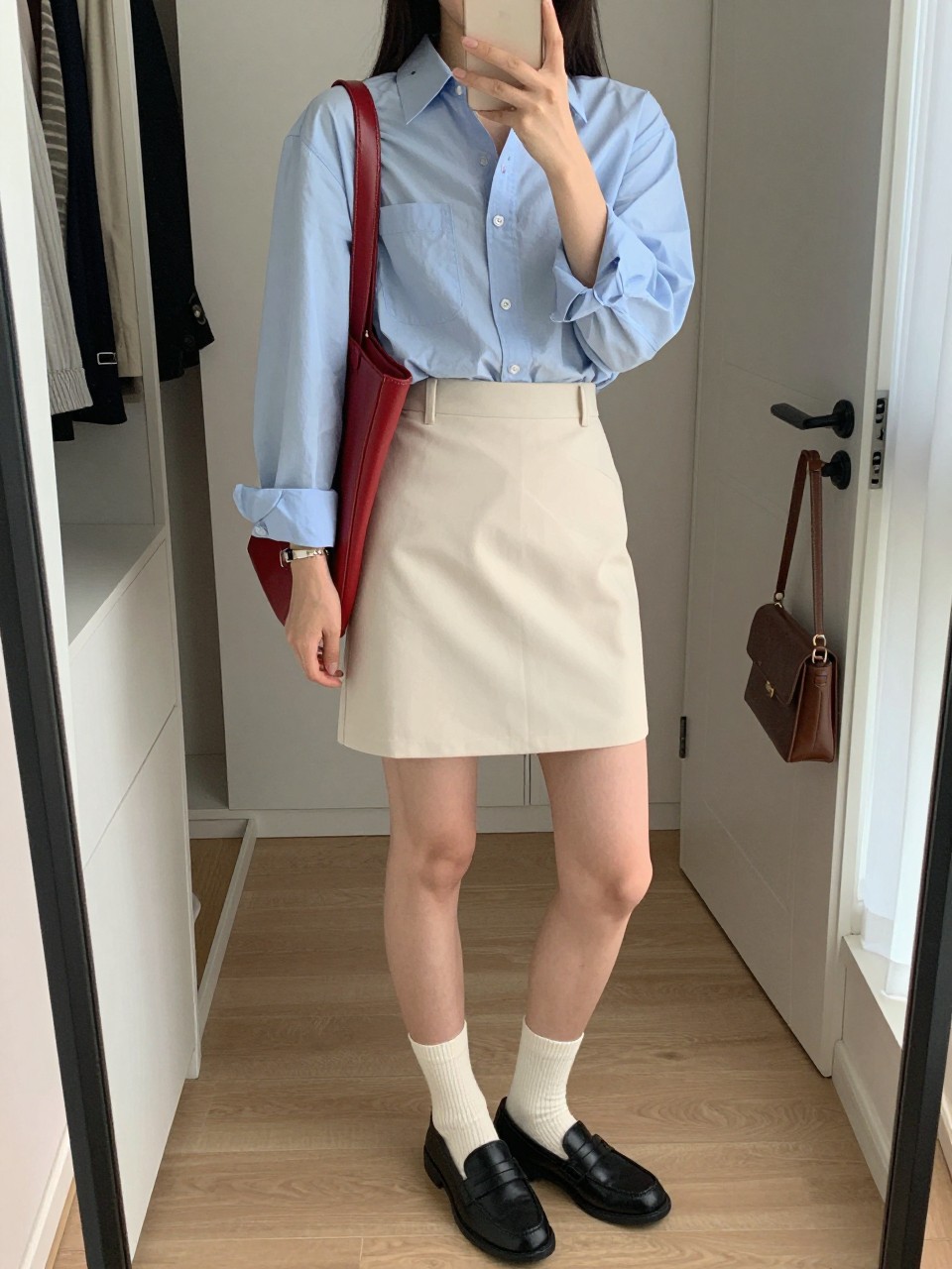 mirror selfie of a woman wearing the exact outfit described above, clearly showing a pale blue Oxford cotton shirt, a cream structured mini skirt, black leather loafers, a red leather shoulder bag, and soft white ribbed cotton socks, holding a phone in front of a mirror, cropped at NECK so the face does not show, with a relaxed casual selfie pose, in a stylish city apartment, natural window light, including open closet with clothes visible and handbag hanging on a door handle, latest iPhone photo quality. (no watermarks on images)