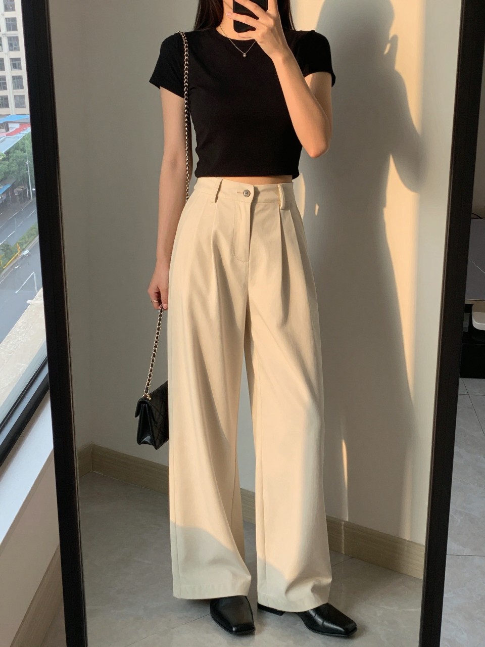 iPhone mirror selfie of a woman in a black cropped top and soft cream wide leg pants with black mules, holding a mini chain bag, hand on hip, cropped at NECK so face doesn't show, in a city apartment, evening golden hour, iPhone photo quality.