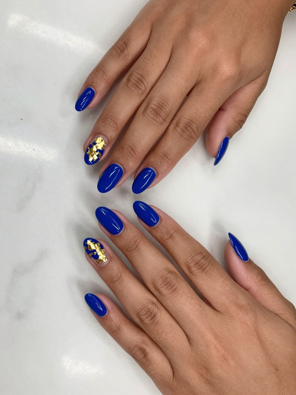 90-degree overhead top-down view of two feminine black hands with almond medium nails.
The manicure features a glossy royal blue base with scattered gold foil accents on two accent nails.
Include reflective metallic detail with a glossy gel finish.

Hands are posed fingers gently spread.

Scene set on a marble surface with a small gold bracelet nearby.

Bright natural daylight with soft shadows, hyper-realistic lifestyle photography, sharp focus on nails, natural skin texture, realistic hands, no extra fingers.