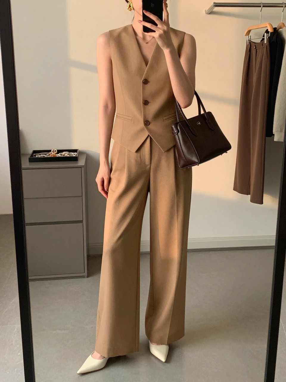mirror selfie of a woman wearing the exact outfit described above, clearly showing a camel tailored vest, matching camel wide-leg tailored trousers, cream pointed mule heels, a dark chocolate leather bag, and gold sculptural earrings, holding a phone in front of a mirror, cropped at NECK so the face does not show, with a relaxed casual selfie pose, in a minimal dressing room, evening golden hour, including jewelry tray on the vanity and clothing rack with garments behind, latest iPhone photo quality. (no watermarks on images)