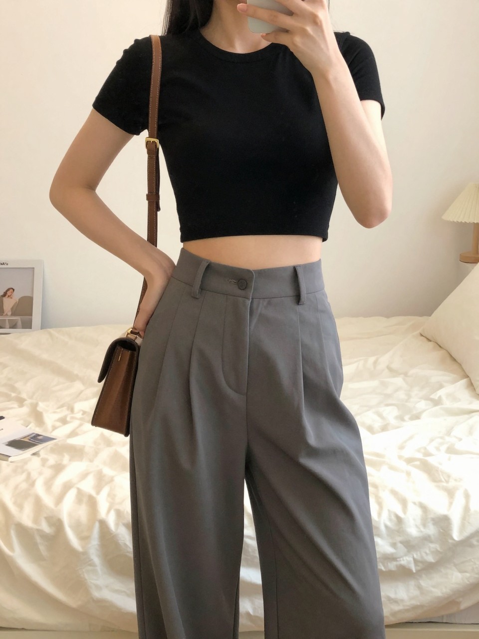 iPhone mirror selfie of a woman in a black cropped top with high-waisted wide leg pants, hand on hip showing the waist definition, cropped at NECK so face doesn't show, in a bright feminine-themed bedroom, warm moody lighting, iPhone photo quality.