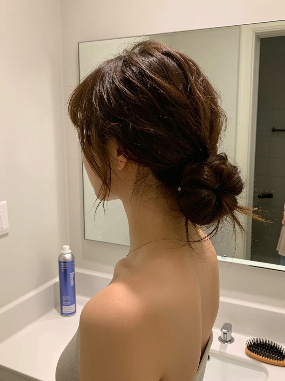 Photo of a woman wearing a loose low bun with soft face-framing pieces, side profile view, setting in a bathroom vanity mirror, lighting soft indoor lighting, containing a small lived-in detail such as a dry shampoo can on the counter and a hairbrush on the vanity, latest iPhone photo quality.