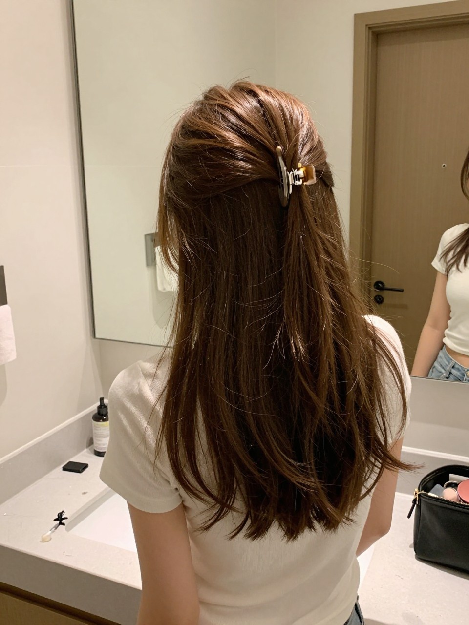 Photo of a woman wearing a claw clip twist on medium-length hair with soft front pieces, back-of-head view, setting in a bathroom vanity mirror, lighting soft indoor lighting, containing a small lived-in detail such as a claw clip on the counter and a makeup bag nearby, latest iPhone photo quality.