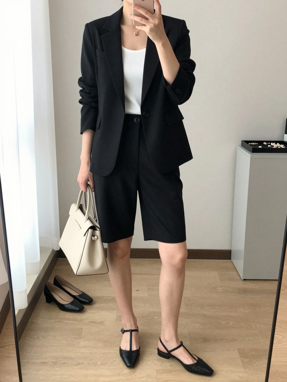 mirror selfie of a woman wearing the exact outfit described above, clearly showing a black tailored blazer, black tailored Bermuda shorts, a white ribbed tank, black strappy low heels, and a structured cream leather bag, holding a phone in front of a mirror, cropped at NECK so the face does not show, with a relaxed casual selfie pose, in a minimal dressing room, natural window light, including pair of heels on the floor nearby and jewelry tray on the vanity, latest iPhone photo quality. (no watermarks on images)