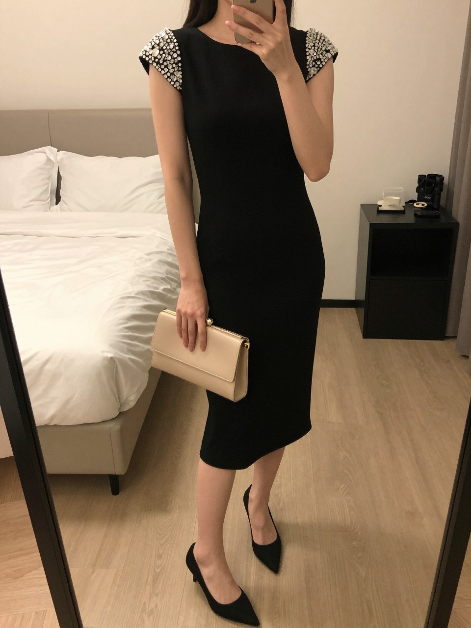 iPhone mirror selfie of a woman wearing a black dress with crystal embellished shoulders and simple black pointed-toe pumps, holding a nude clutch, posed to highlight the shoulder detail, cropped at NECK so face doesn't show, in a feminine-themed bedroom, warm moody lighting, iPhone photo quality.