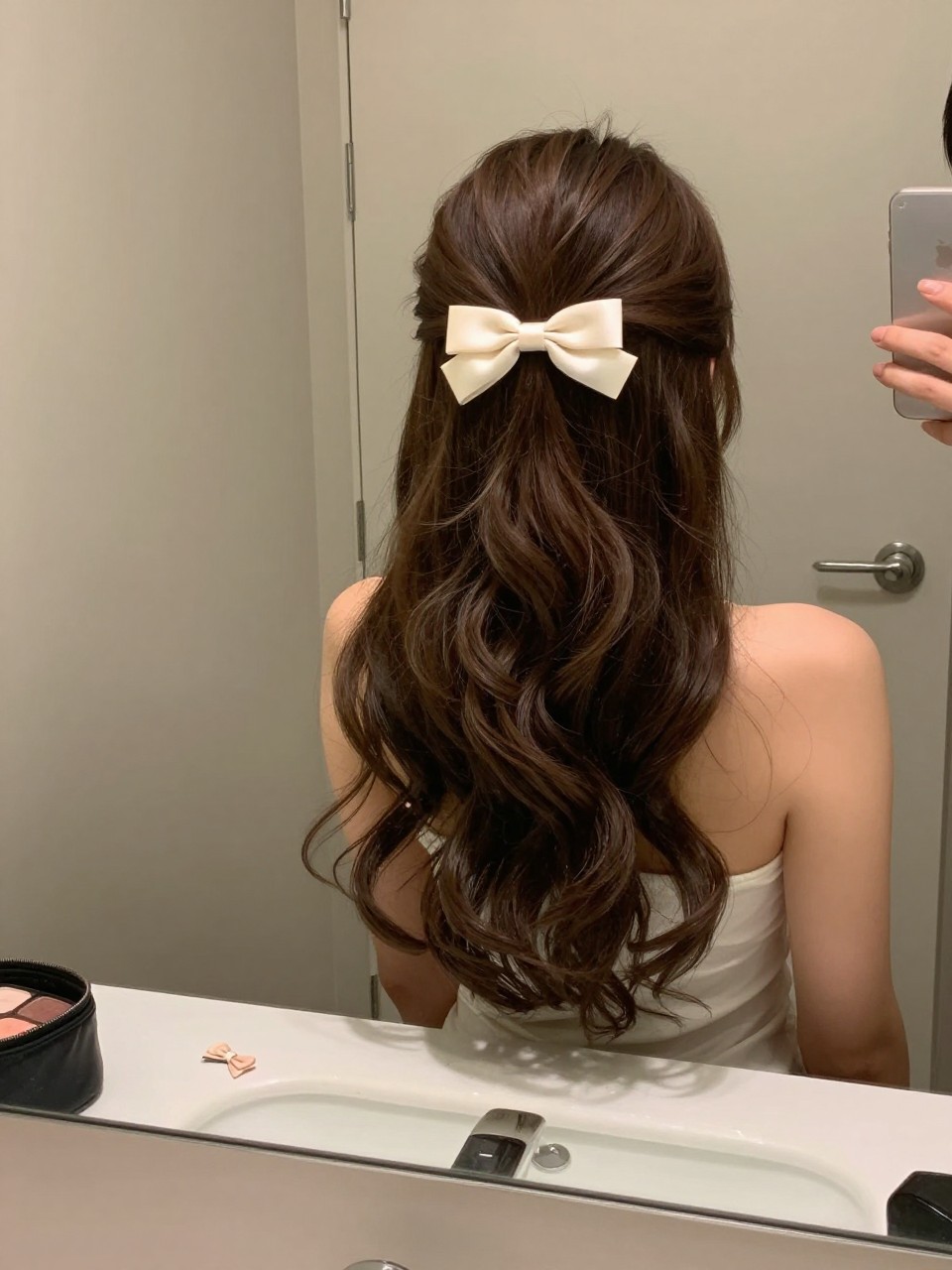 Photo of a woman wearing a half up half down hairstyle with a satin bow clip and soft curls down, candid mirror selfie, setting in a bathroom vanity mirror, lighting soft indoor lighting, containing a small lived-in detail such as a small bow clip on the counter and a makeup bag nearby, latest iPhone photo quality.