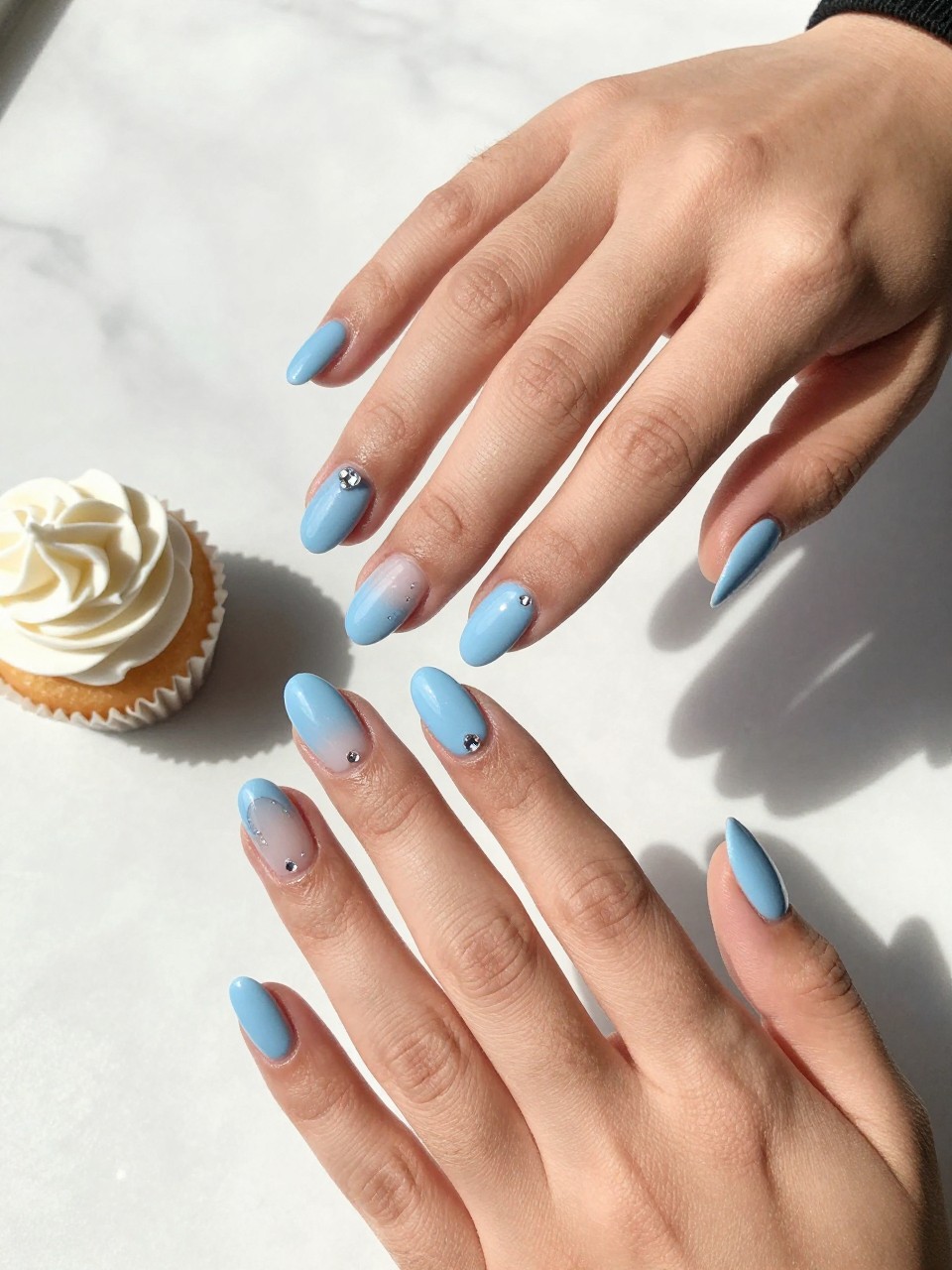 90-degree overhead top-down view of two feminine hands with almond medium nails.
The manicure features a glossy baby blue base with tiny crystal accents placed near the cuticle on two accent nails.
Include reflective gemstone details with a glossy gel finish.

Hands are posed with fingers gently spread.

Scene set on a white marble surface with a small birthday cupcake nearby.

Bright natural daylight with soft shadows, hyper-realistic lifestyle photography, sharp focus on nails, natural skin texture, realistic hands, no extra fingers.