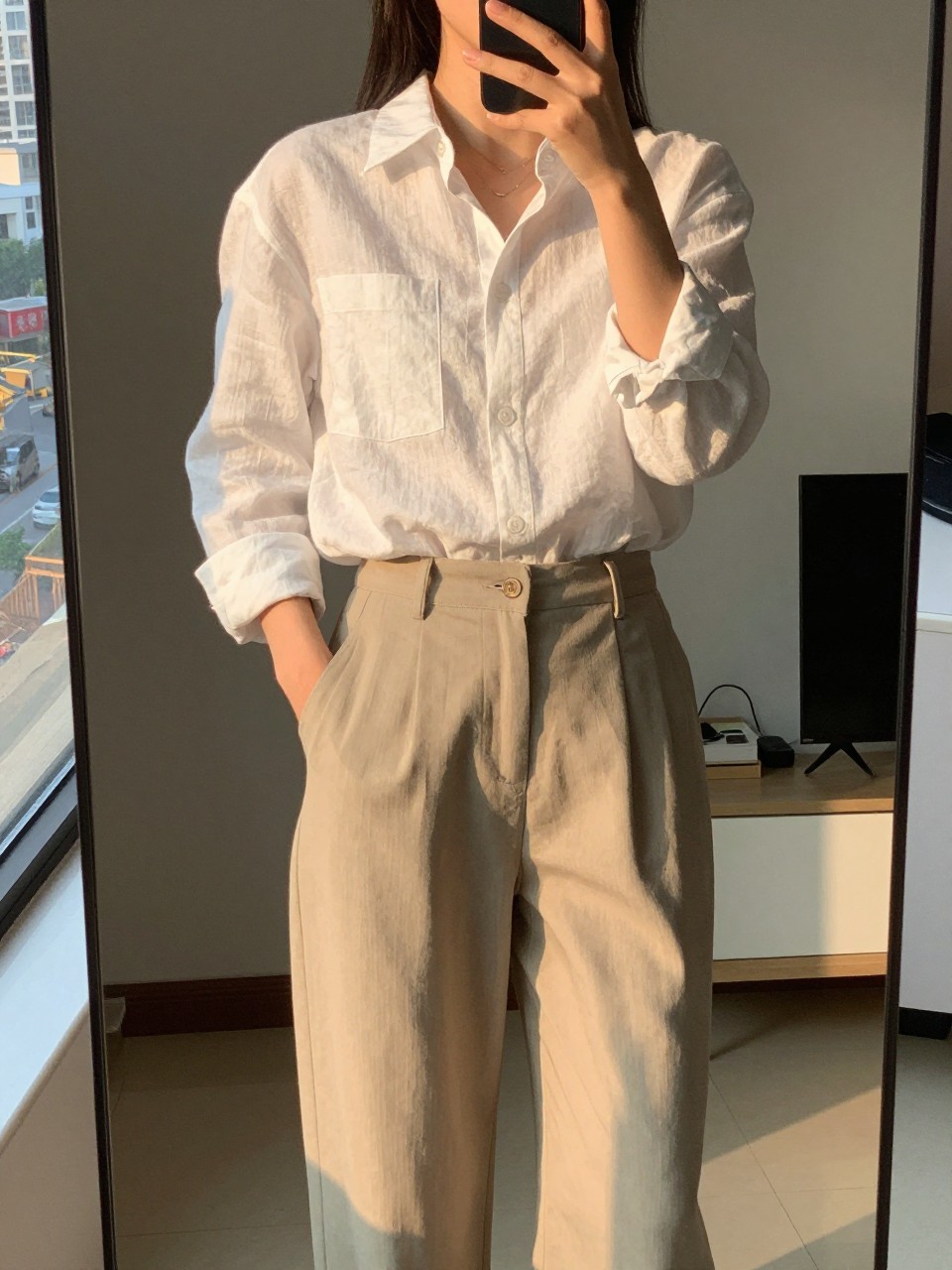iPhone mirror selfie of a woman wearing a crisp white linen shirt tucked into flowy beige trousers, standing with one hand in her pocket and looking slightly to the side, cropped at NECK so face doesn't show, in a sunlit city apartment, evening golden hour, iPhone photo quality.