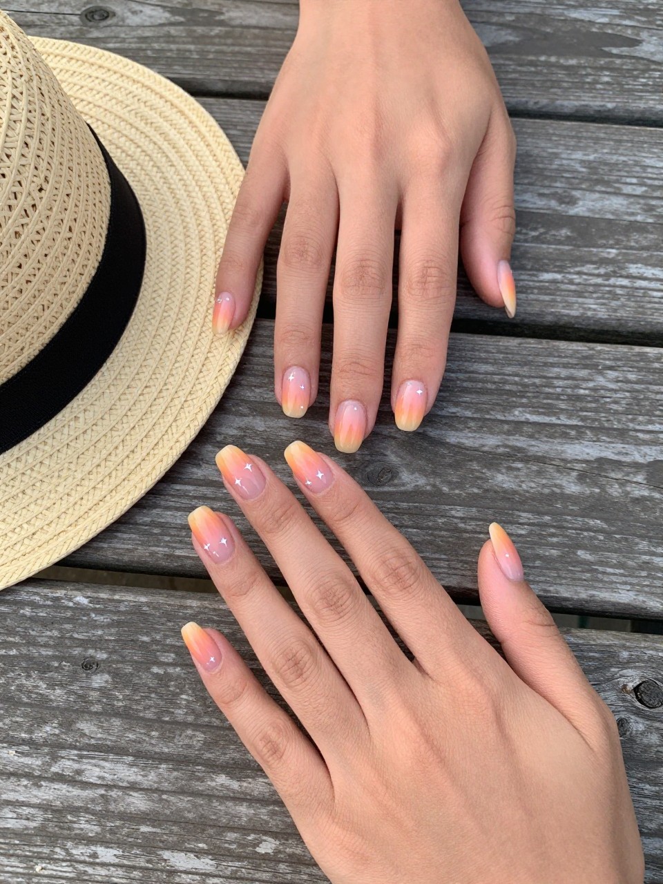 90-degree overhead top-down view of two feminine black hands with almond medium nails.
The manicure features a peach to orange sunset ombré base with tiny white star accents on two nails.
Include glossy gel finish.

Hands are posed fingers gently spread.

Scene set on a wooden picnic table with a small straw hat nearby.

Bright natural daylight with soft shadows, hyper-realistic lifestyle photography, sharp focus on nails, natural skin texture, realistic hands, no extra fingers.