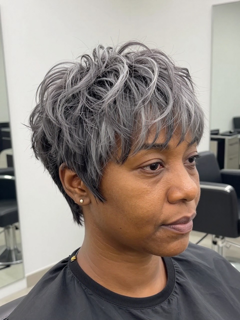 Photo of a black woman with grey hair wearing a soft coily taper cut with neatly shaped sides and fuller defined height on top, with the haircut fully visible and clearly defined, shot from a front portrait view, in a bright salon interior, with soft indoor lighting, showing the haircut shape, layers, ends, bangs, and texture clearly, including simple gold earrings, latest iPhone photo quality.