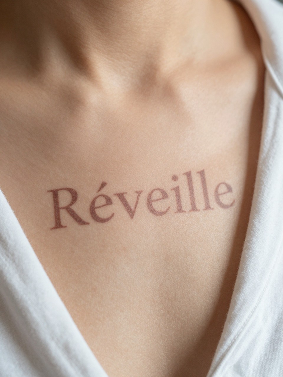 "Extreme close-up of 'Réveille' on the ribcage, the letters seeming to blur into clarity. A simple, white cotton sleep shirt is visible. Soft, dawn-like lighting on the skin."