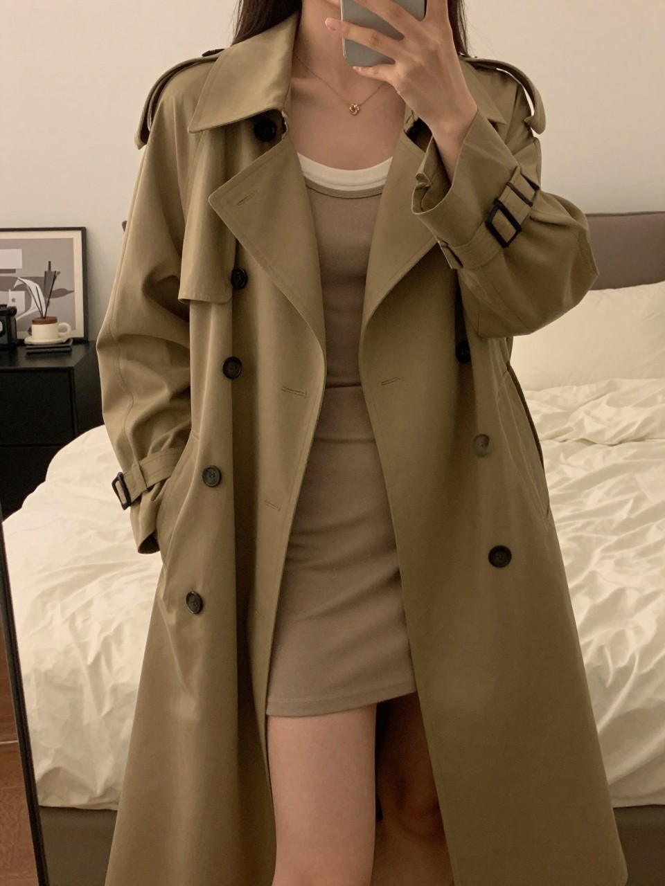 iPhone mirror selfie of a woman wearing an open trench coat over a t-shirt and slip skirt, holding the lapel of the coat with one hand, cropped at NECK so face doesn't show, in a feminine-themed bedroom, warm moody lighting, iPhone photo quality.