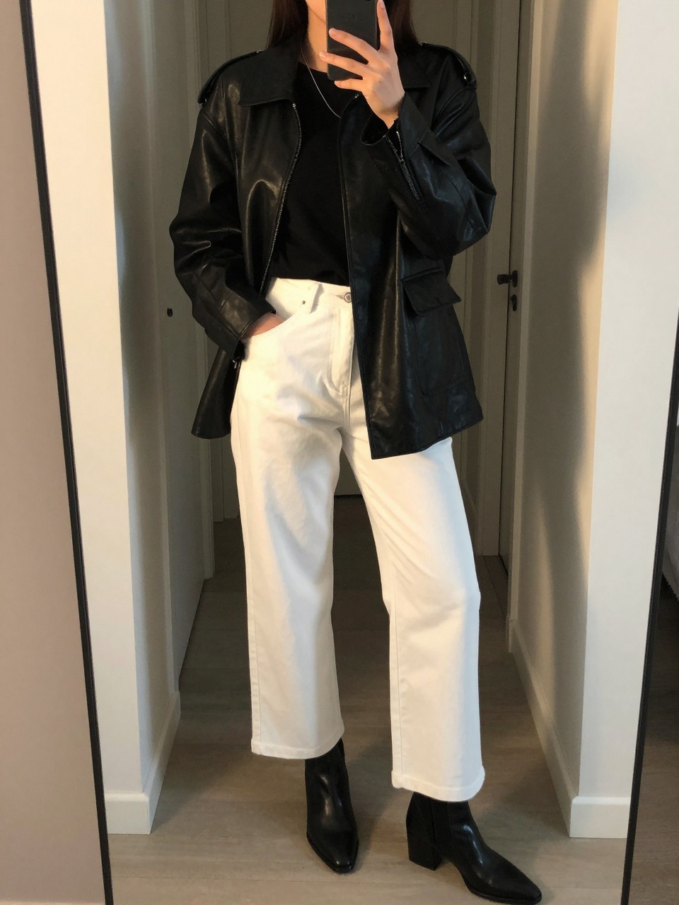 iPhone mirror selfie of a woman in a black leather jacket over a tee with white wide leg pants and ankle boots, one hand in jacket pocket, cropped at NECK so face doesn't show, in a city apartment entryway, evening golden hour, iPhone photo quality.