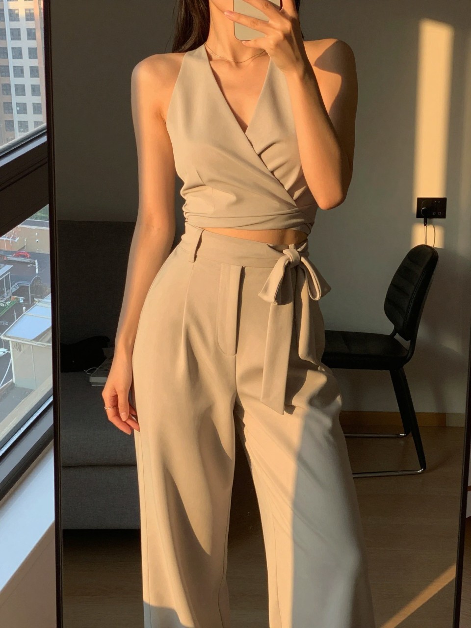 iPhone mirror selfie of a woman in a wrap top tucked into wide leg pants, showing the tied waist detail, standing with slight lean, cropped at NECK so face doesn't show, in a city apartment, evening golden hour, iPhone photo quality.