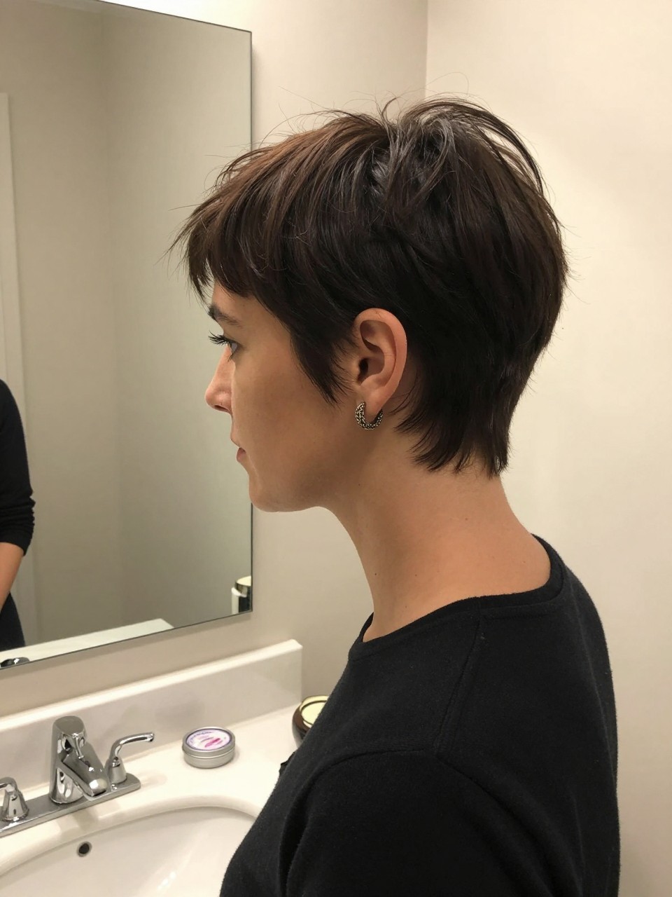 Photo of a BRUNNETE woman wearing a polished pixie with a deep side part, side profile view, setting in a bathroom vanity mirror, lighting soft indoor lighting, containing a small lived-in detail such as a small pomade tin on the counter and statement earrings peeking through, latest iPhone photo quality.