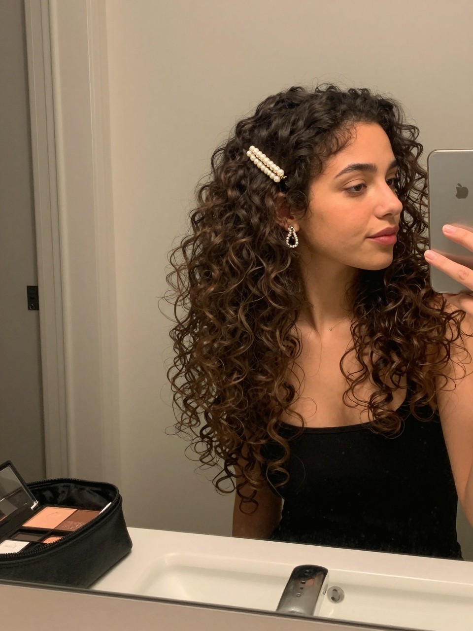 Photo of a woman wearing loose curls half up half down secured with a pearl barrette, candid mirror selfie, setting in a bathroom vanity mirror, lighting soft indoor lighting, containing a small lived-in detail such as a makeup bag nearby and statement earrings peeking through, latest iPhone photo quality.