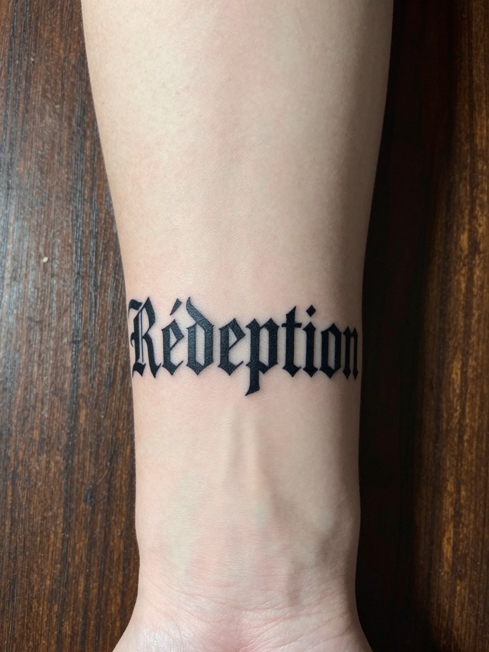 "Close-up of 'Rédemption' in strong, Gothic-inspired script on the forearm. The arm is against a backdrop of rough-hewn, but polished, dark wood. Serious and redeemed."