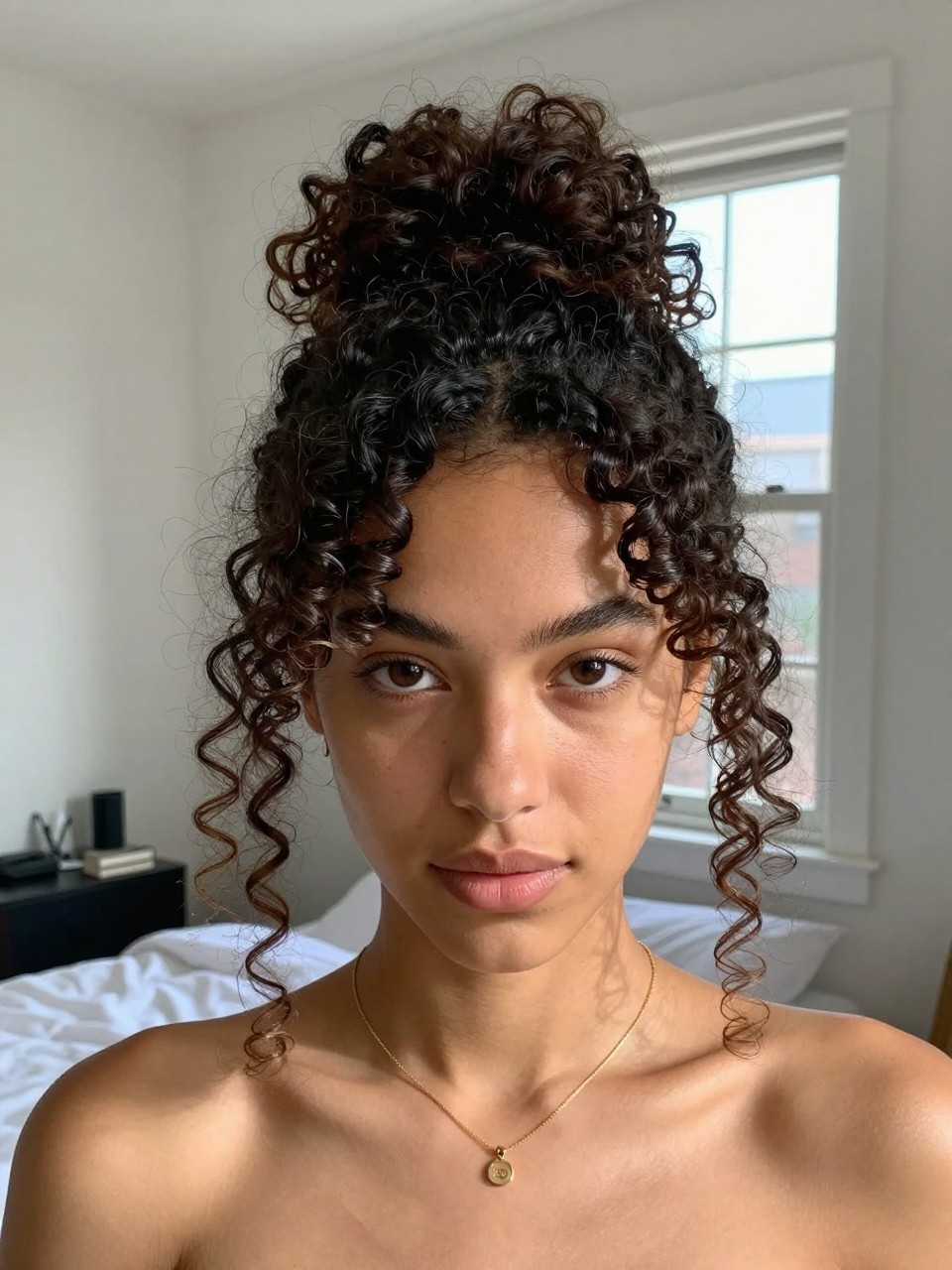 Photo of a woman wearing a curly top knot with face-framing pieces, front-facing view showing loose curls around the face, setting is a sun-drenched bedroom with natural window light, containing a simple gold necklace visible, iPhone photo quality.