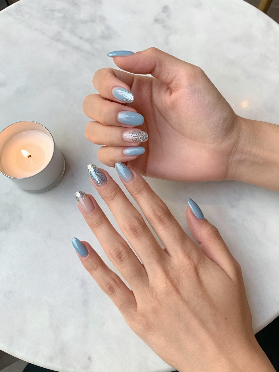 90-degree overhead top-down view of two feminine hands with almond medium nails.
The manicure features a soft blue base with silver glitter concentrated along the tips.
Include reflective sparkle with a glossy gel finish.

Hands are posed one hand resting over the other.

Scene set on a marble café table with a small birthday candle nearby.

Bright natural daylight with soft shadows, hyper-realistic lifestyle photography, sharp focus on nails, natural skin texture, realistic hands, no extra fingers.