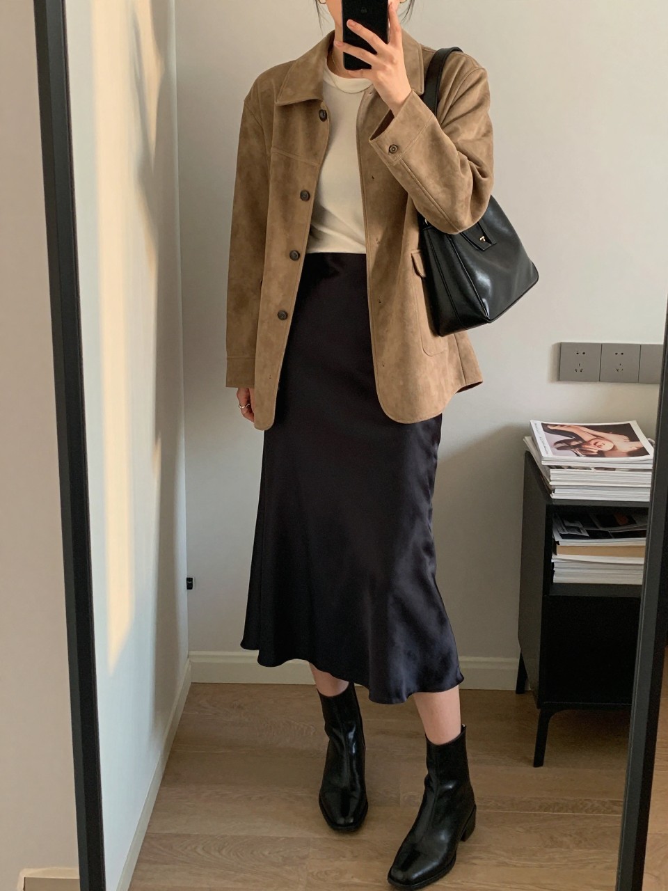 mirror selfie of a woman wearing the exact outfit described above, clearly showing a soft tan suede jacket, a black satin slip midi skirt, an ivory fitted tee, black ankle leather boots, and a structured black shoulder bag, holding a phone in front of a mirror, cropped at NECK so the face does not show, with a relaxed casual selfie pose, in a stylish city apartment, evening golden hour, including handbag hanging on a door handle and stack of fashion magazines on a side table, latest iPhone photo quality. (no watermarks on images)