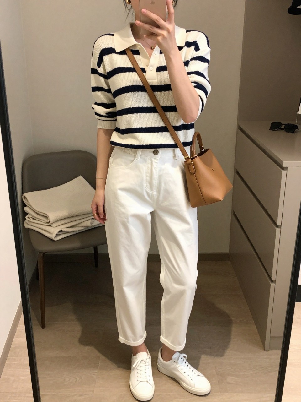 mirror selfie of a woman wearing the exact outfit described above, clearly showing a navy and cream striped polo knit top, white barrel-leg cotton twill pants, clean white leather sneakers, a soft tan leather crossbody bag, and small gold hoop earrings, holding a phone in front of a mirror, cropped at NECK so the face does not show, with a relaxed casual selfie pose, in a walk-in closet mirror, soft indoor lighting, including folded blanket on a chair and sunglasses placed on a dresser, latest iPhone photo quality. (no watermarks on images)