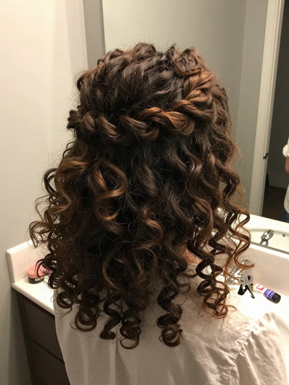 Photo of a woman wearing a half-up twisted crown on naturally curly hair with defined curls down, back-of-head view to highlight the twist detail, setting in a bathroom vanity mirror, lighting soft indoor lighting, containing a small lived-in detail such as a curl cream tube on the counter and a hair clip clipped to the collar, latest iPhone photo quality.