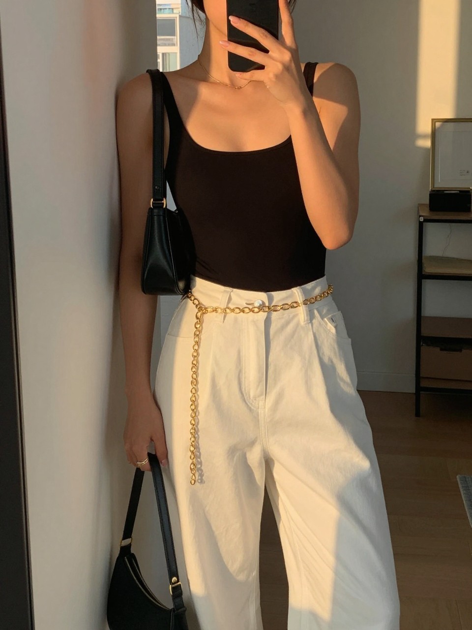 iPhone mirror selfie of a woman in a black bodysuit tucked into white wide leg pants with a gold chain belt, holding a black shoulder bag, relaxed lean against a wall, cropped at NECK so face doesn't show, in a city apartment, evening golden hour, iPhone photo quality.