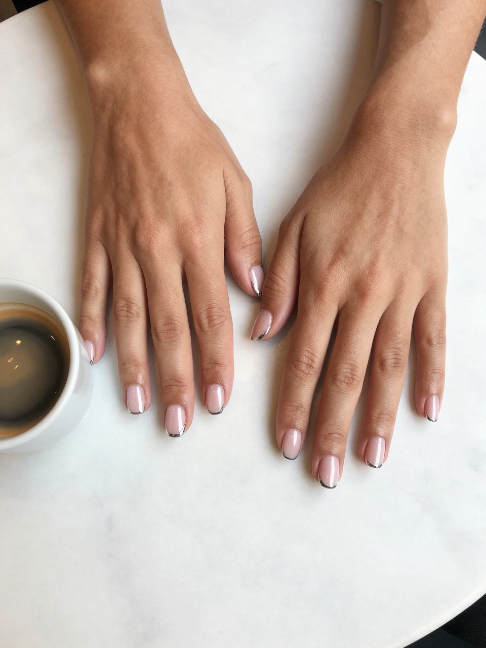 90-degree overhead top-down view of two feminine black hands with square short nails.
The manicure features a milky pink chrome base with thin silver chrome French tips.
Include metallic reflective shine with a glossy gel finish.

Hands are posed resting flat.

Scene set on a white marble café table with a small espresso cup nearby.

Bright natural daylight with soft shadows, hyper-realistic lifestyle photography, sharp focus on nails, natural skin texture, realistic hands, no extra fingers.