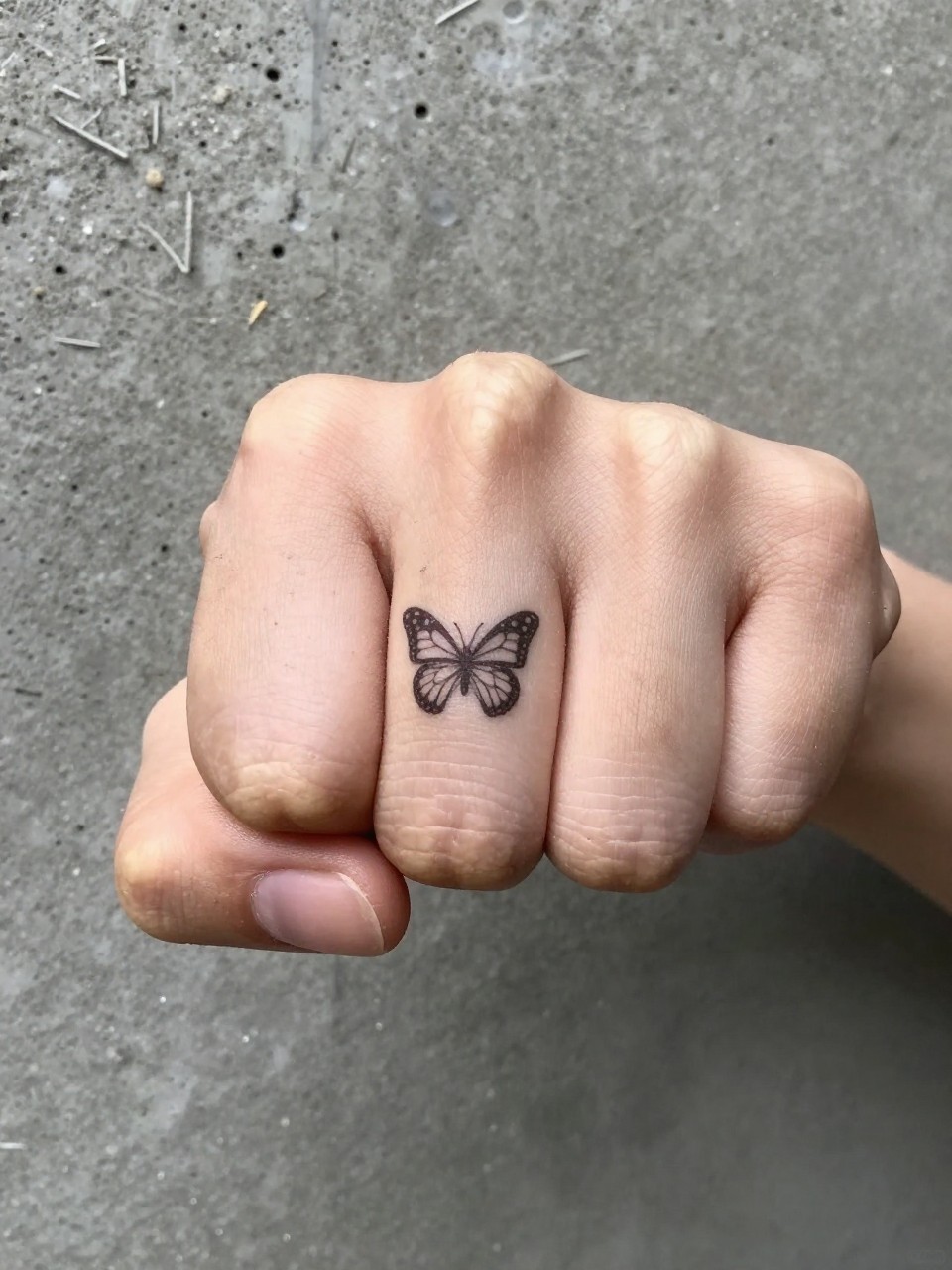 "Close-up of a clenched fist: micro butterfly silhouettes on each knuckle. Shot against a concrete wall, highlighting the contrast between delicate symbol and tough placement."