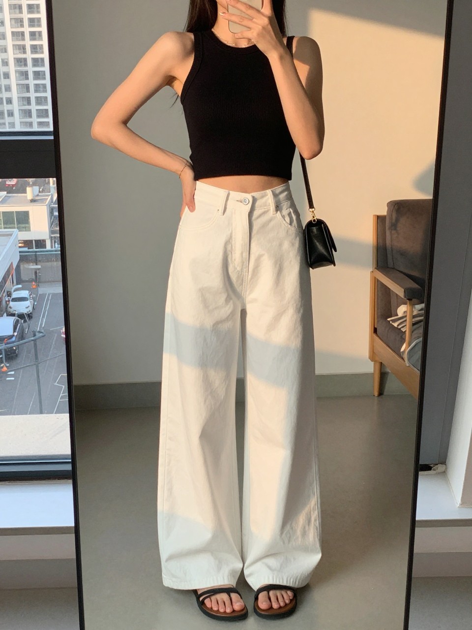 iPhone mirror selfie of a woman in a black ribbed crop tank with white wide leg pants and slide sandals, holding a tiny shoulder bag, hand on hip, cropped at NECK so face doesn't show, in a city apartment, evening golden hour, iPhone photo quality.