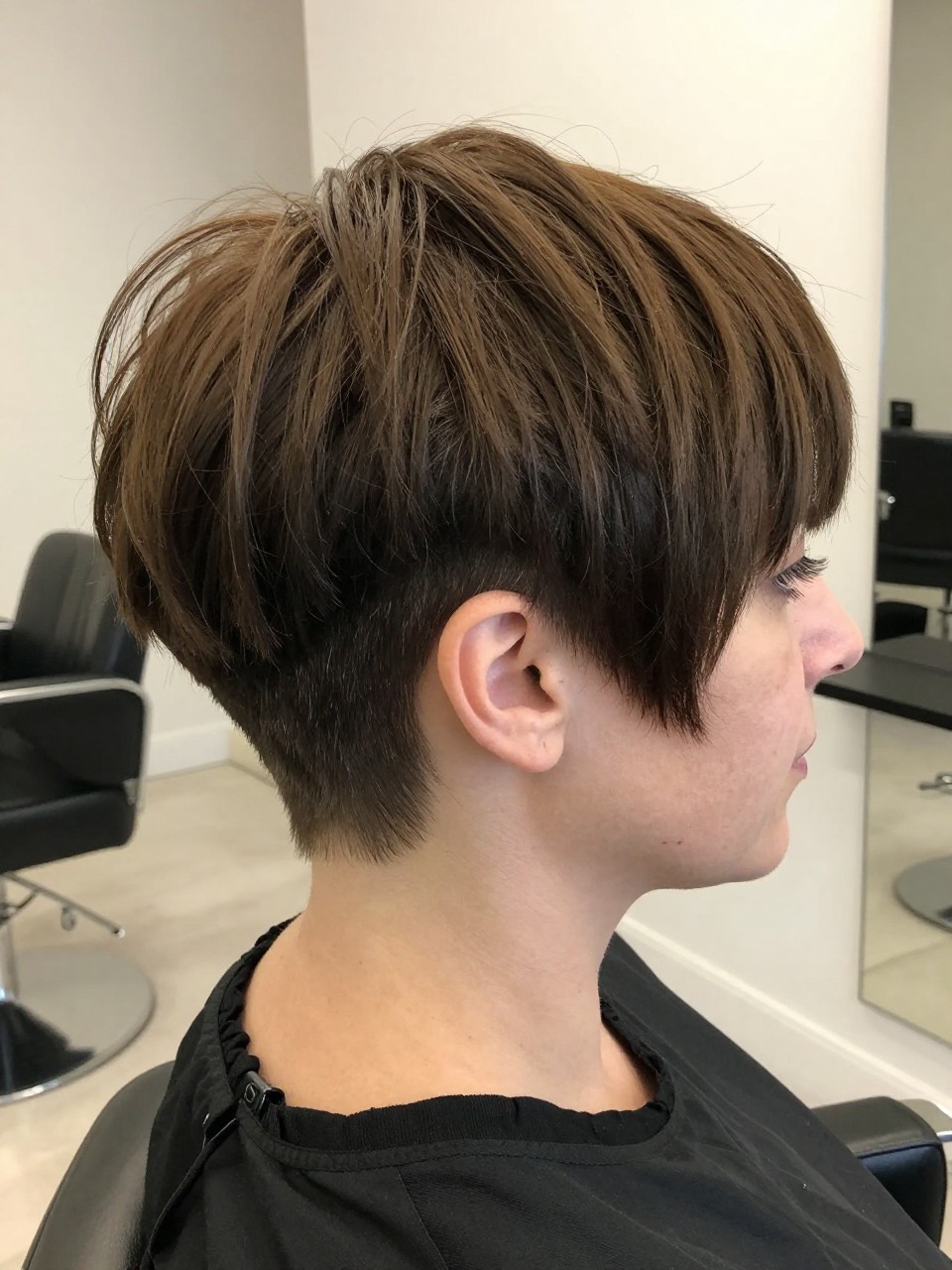 Photo of a white woman wearing an ultra short undercut pixie with very close sides and a slightly longer shaped top, with the haircut fully visible and clearly defined, shot from a side profile, in a bright salon interior, with soft indoor lighting, showing the haircut shape, layers, ends, bangs, and texture clearly, including the salon chair edge, latest iPhone photo quality.