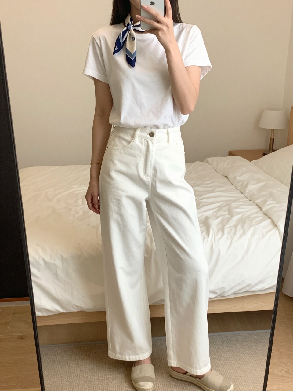 iPhone mirror selfie of a woman in a simple tee with a silk scarf at neck, white wide leg pants and espadrilles, relaxed pose holding phone, cropped at NECK so face doesn't show, in a bright feminine-themed bedroom, warm moody lighting, iPhone photo quality.