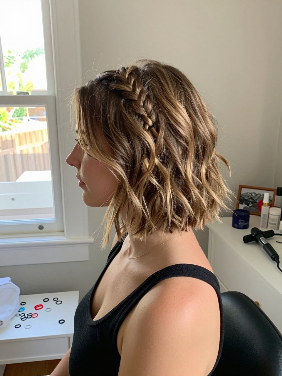 Photo of a white woman wearing a short lob with a small side braid accent and loose waves, side profile view, setting in a sun-drenched bedroom, lighting natural window light, containing a small lived-in detail such as mini elastics on the vanity and a curling iron on the counter, latest iPhone photo quality.