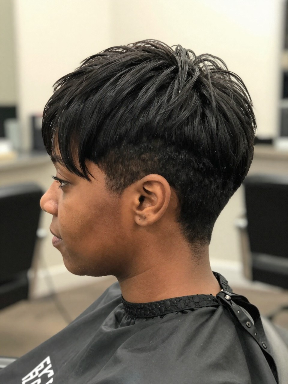 Photo of a black woman wearing an ultra short buzzed taper pixie with very close sides and slightly longer top length, with the haircut fully visible and clearly defined, shot from a side profile, in a bright salon interior, with soft indoor lighting, showing the haircut shape, layers, ends, bangs, and texture clearly, including the salon chair edge, latest iPhone photo quality.