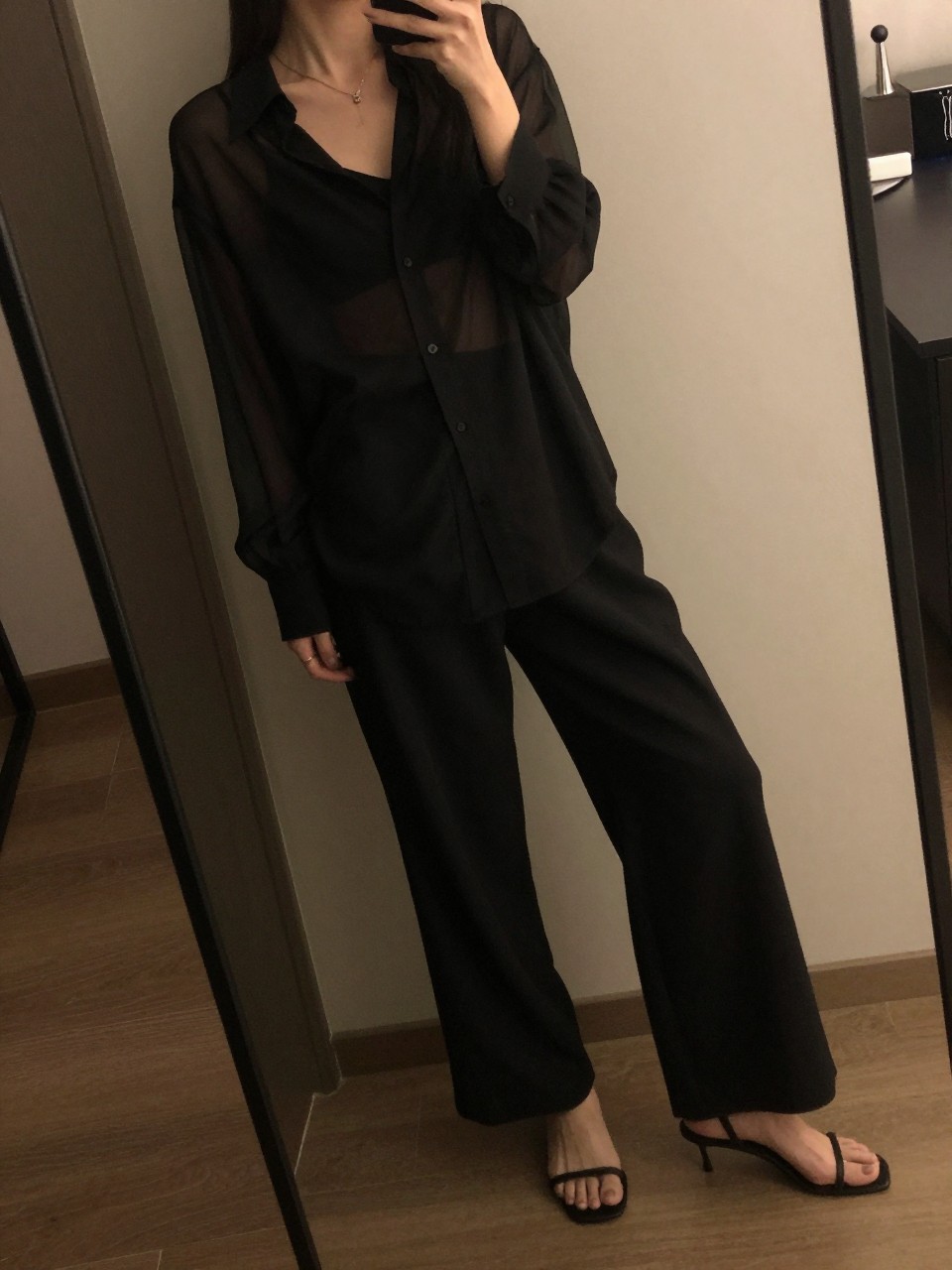 iPhone mirror selfie of a woman in a sheer black blouse over a camisole with wide leg pants and heeled mules, angled pose, cropped at NECK so face doesn't show, in a dim city apartment, warm moody lighting, iPhone photo quality.