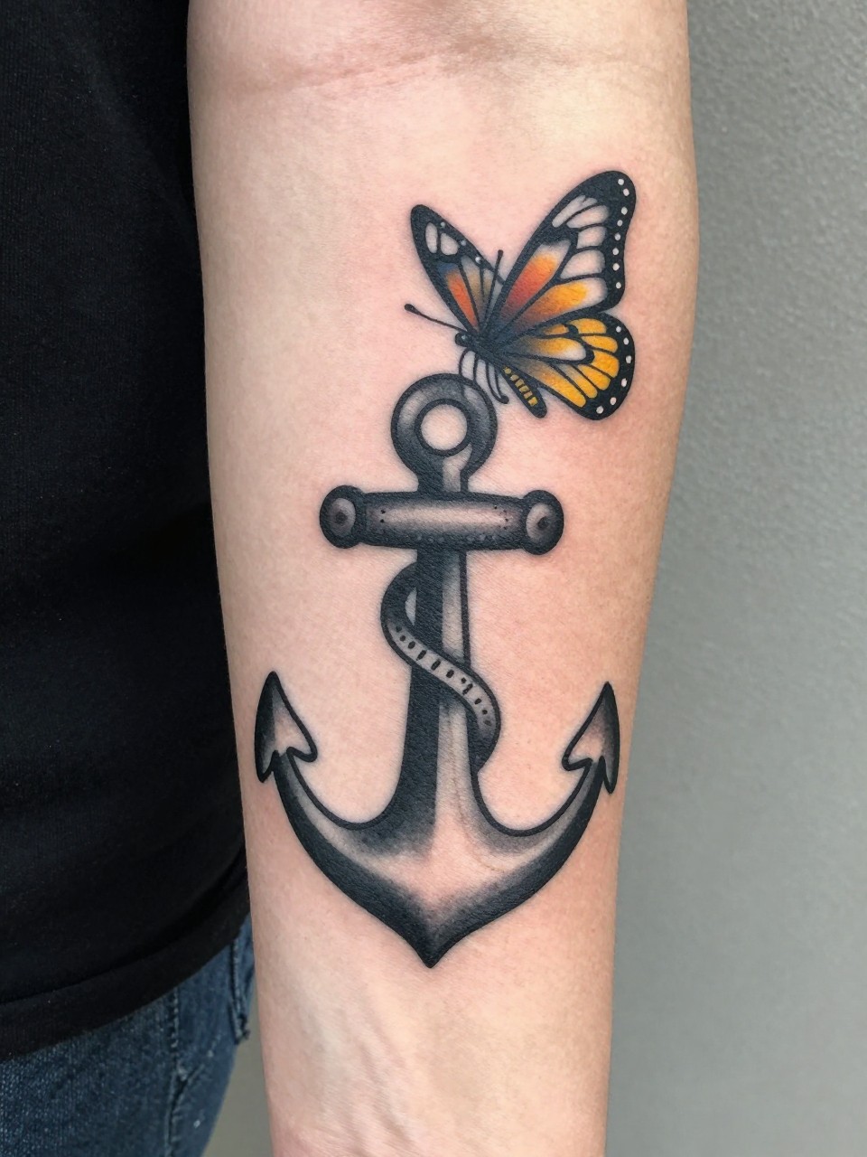 "Inner forearm tattoo: a bold traditional anchor with a single butterfly rising from its top. Black and grey with pops of yellow on the butterfly."