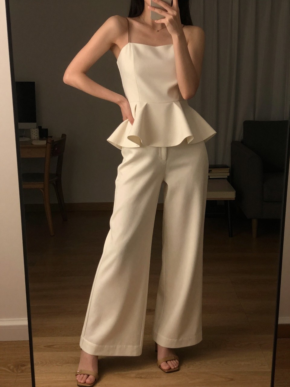 iPhone mirror selfie of a woman in a peplum top with wide leg pants and heeled sandals, hand on hip showing the peplum shape, cropped at NECK so face doesn't show, in a dim city apartment, warm moody lighting, iPhone photo quality.
