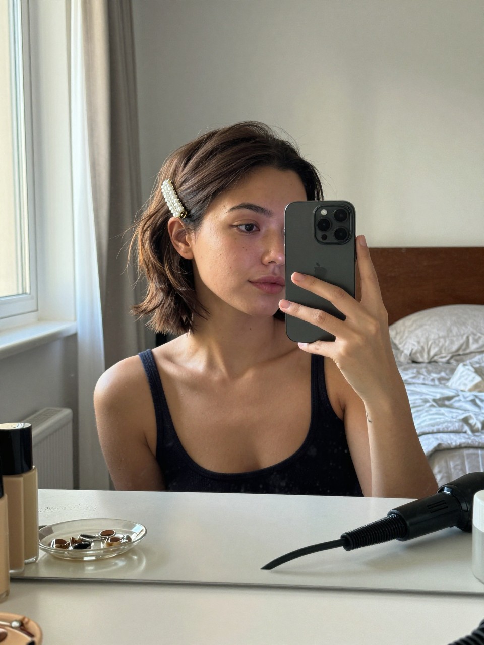 Photo of a woman wearing a half-up twist on short hair secured with a pearl clip, candid mirror selfie, setting in a sun-drenched bedroom, lighting natural window light, containing a small lived-in detail such as a jewelry dish on the counter and a curling iron on the vanity, latest iPhone photo quality.
