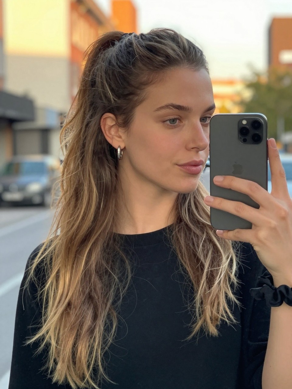 Photo of a white woman wearing a loose half-up ponytail with a lifted crown and the rest of the hair down, candid mirror selfie, setting in a softly blurred city street background, lighting golden hour glow, containing a small lived-in detail such as statement earrings peeking through and a scrunchie on the wrist, latest iPhone photo quality.