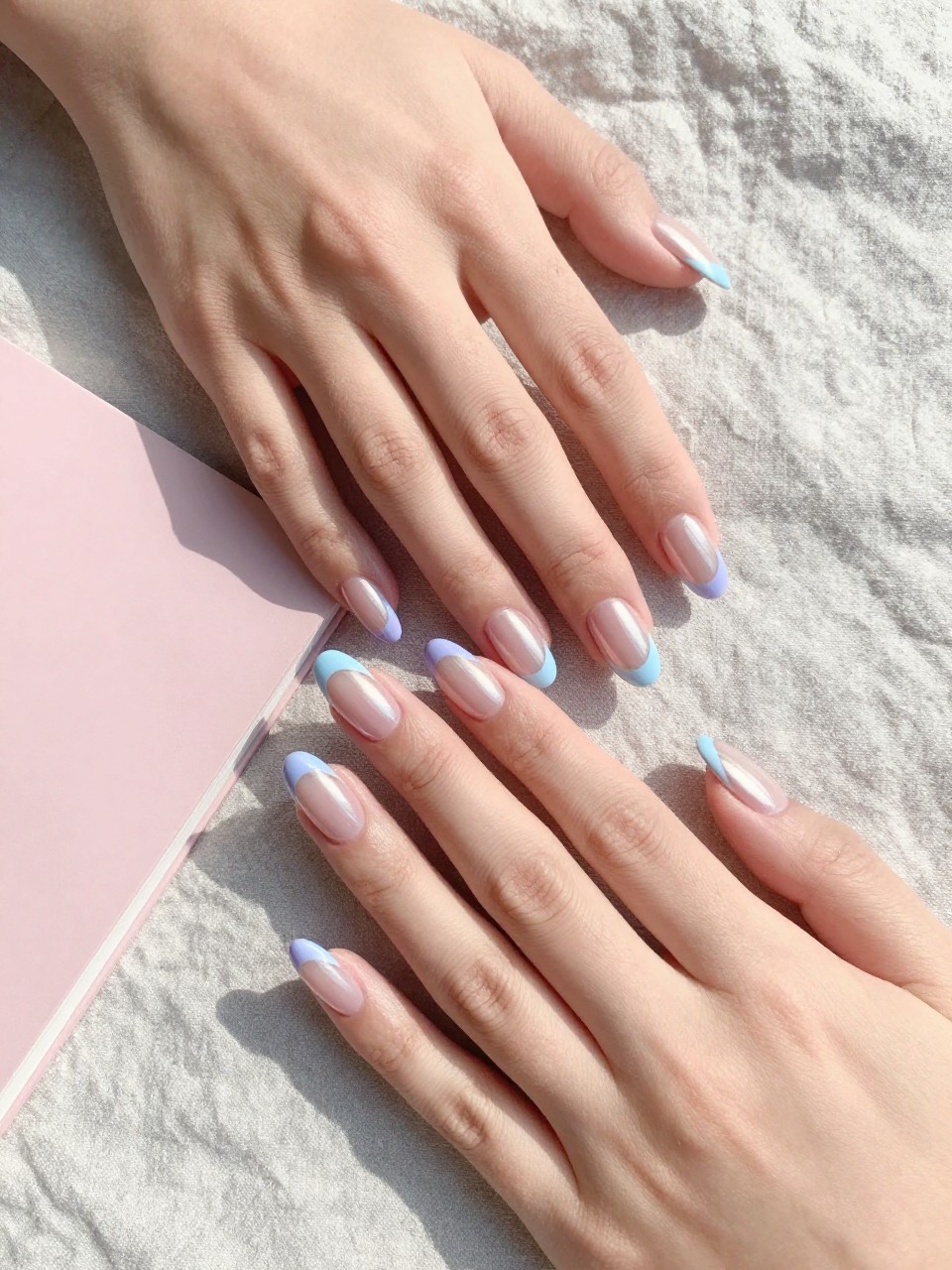 90-degree overhead top-down view of two feminine hands with almond medium nails.
The manicure features a milky pink chrome base with soft pastel French tips in light lavender and baby blue.
Include glossy reflective chrome shine with a gel finish.

Hands are posed one hand placed gently over the other.

Scene set on a soft linen cloth with a small pastel notebook nearby.

Bright natural daylight with soft shadows, hyper-realistic lifestyle photography, sharp focus on nails, natural skin texture, realistic hands, no extra fingers.