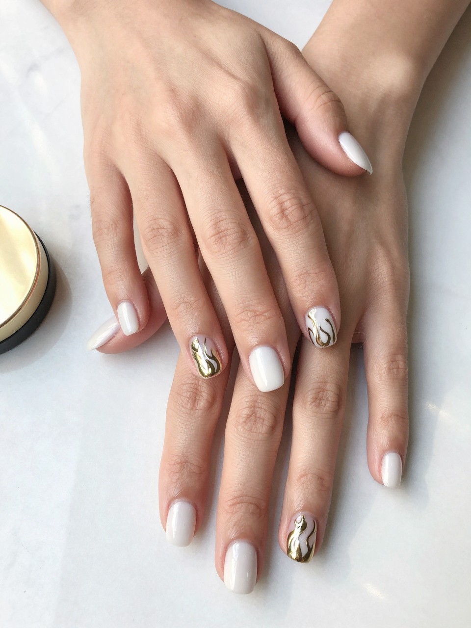 90-degree overhead top-down view of two feminine hands with rounded square medium nails.
The manicure features a glossy white base with metallic gold flame details painted across three accent nails.
Include reflective chrome-like accents with a glossy gel finish.

Hands are posed resting flat.

Scene set on a white marble surface with a small gold compact nearby.

Bright natural daylight with soft shadows, hyper-realistic lifestyle photography, sharp focus on nails, natural skin texture, realistic hands, no extra fingers.