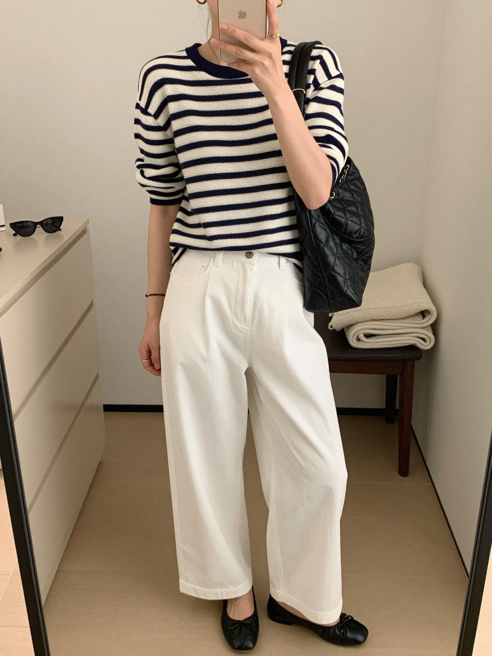 mirror selfie of a woman wearing the exact outfit described above, clearly showing a navy and cream fine knit striped top, white wide-leg trousers, black leather ballet flats, a quilted black leather shoulder bag, and small gold hoop earrings, holding a phone in front of a mirror, cropped at NECK so the face does not show, with a relaxed casual selfie pose, in a walk-in closet mirror, soft indoor lighting, including sunglasses placed on a dresser and folded blanket on a chair, latest iPhone photo quality. (no watermarks on images)