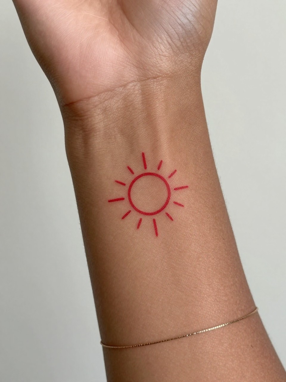 "design-focused close-up of a tiny red sun tattoo on deep brown skin at the wrist, clean red ink, small circle with thin rays, minimalist style, realistic skin texture, delicate bracelet, soft minimal background, modern feminine tattoo inspiration photography (no watermarks on images)"
