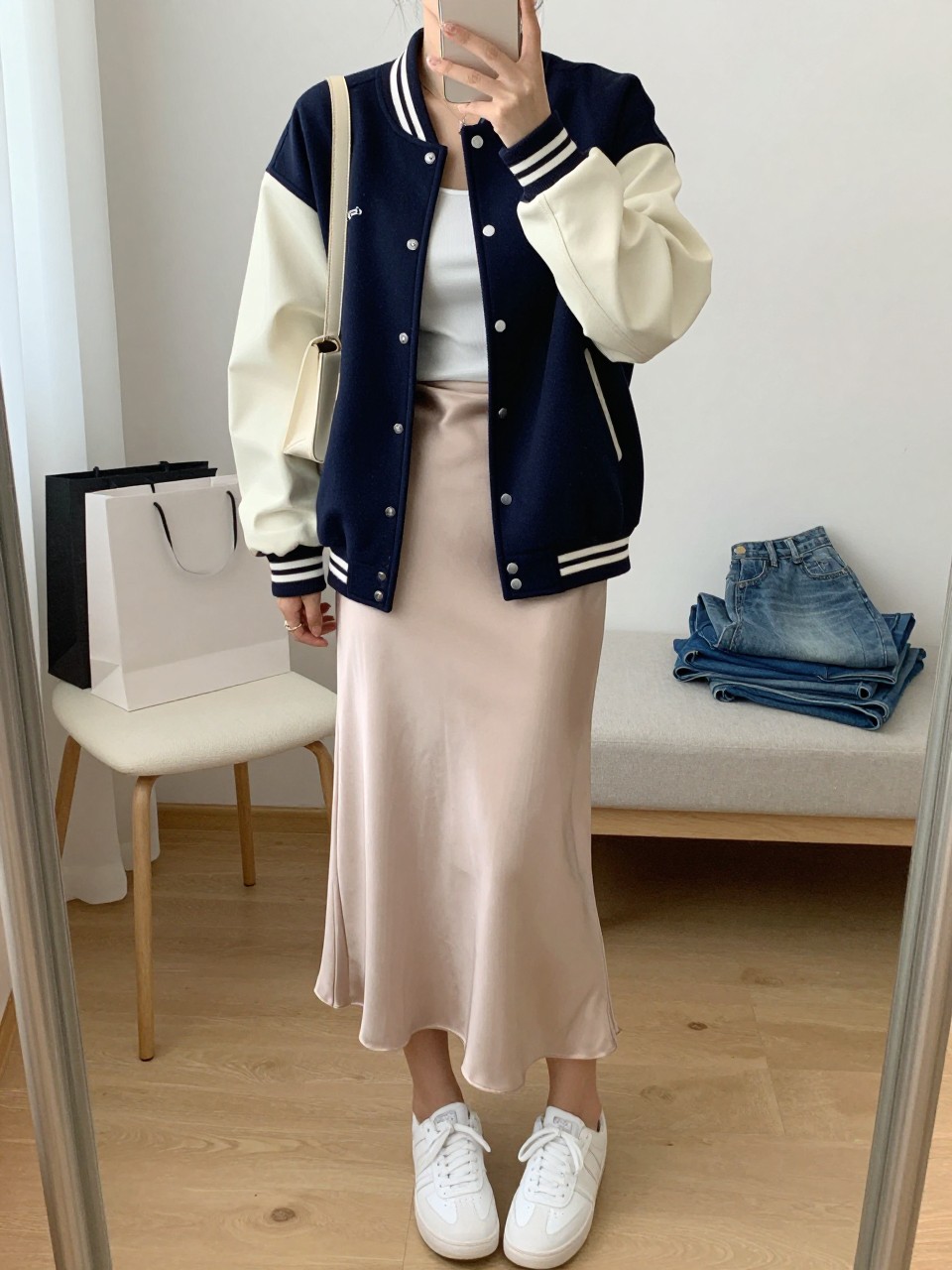 mirror selfie of a woman wearing a navy and cream varsity jacket with a structured fit, a champagne satin column midi skirt, a white fitted tank top, white clean sneakers, and a cream shoulder bag, holding a phone in front of a mirror, cropped at NECK so the face does not show, with a relaxed casual selfie pose, in a feminine-themed bedroom, natural window light, including shopping bags beside a chair and folded denim jeans on a bench, latest iPhone photo quality. (no watermarks on images)