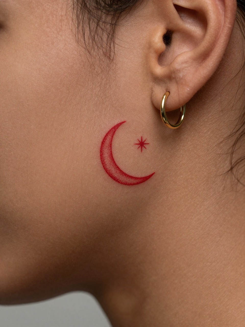  "macro shot of a red crescent moon and tiny star tattoo on rich brown skin behind the ear, clean red ink, fine line celestial details, minimalist style, realistic skin texture, small gold hoop earring, tight crop, soft blurred background, design-first tattoo inspiration image (no watermarks on images)"