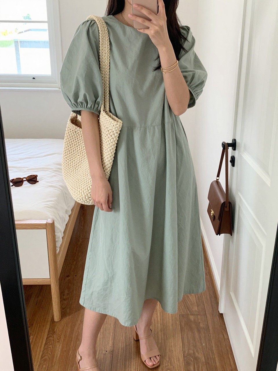mirror selfie of a woman wearing the exact outfit described above, clearly showing a soft sage cotton midi dress with puff sleeves, nude leather block-heel sandals, a woven cream-texture shoulder bag, soft tortoise shell sunglasses, and a thin gold bracelet stack, holding a phone in front of a mirror, cropped at NECK so the face does not show, with a relaxed casual selfie pose, in a feminine-themed bedroom, natural window light, including sunglasses placed on a dresser and handbag hanging on a door handle, latest iPhone photo quality. (no watermarks on images)