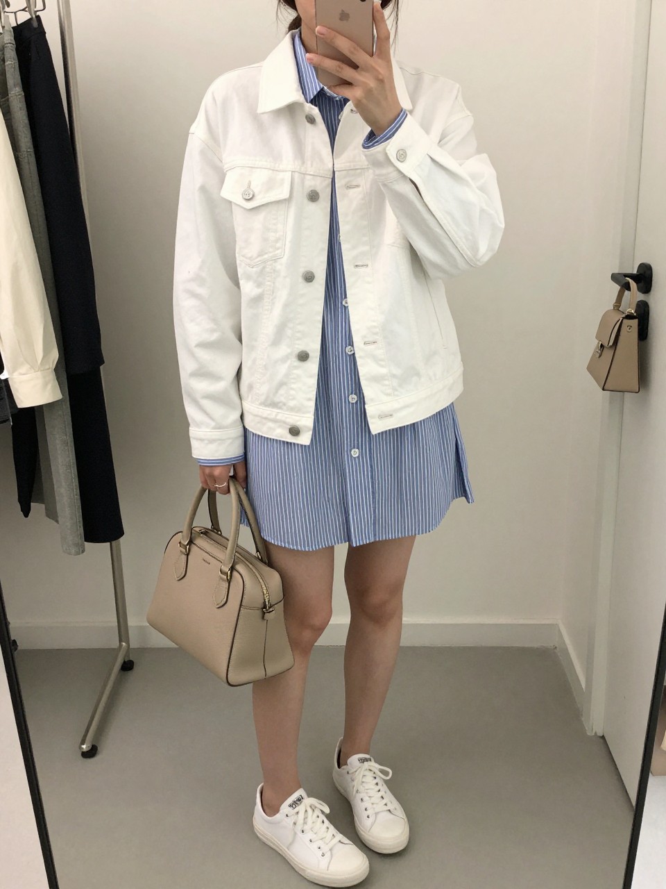 mirror selfie of a woman wearing the exact outfit described above, clearly showing a white denim jean jacket with silver buttons, blue and white striped shirt dress, white low-top sneakers, light tan structured handbag, and a low sleek ponytail, holding a phone in front of a mirror, cropped at NECK so the face does not show, with a relaxed casual selfie pose, in a fashion studio mirror corner, soft indoor lighting, including a clothing rack with garments behind and a handbag hanging on a door handle, latest iPhone photo quality.
