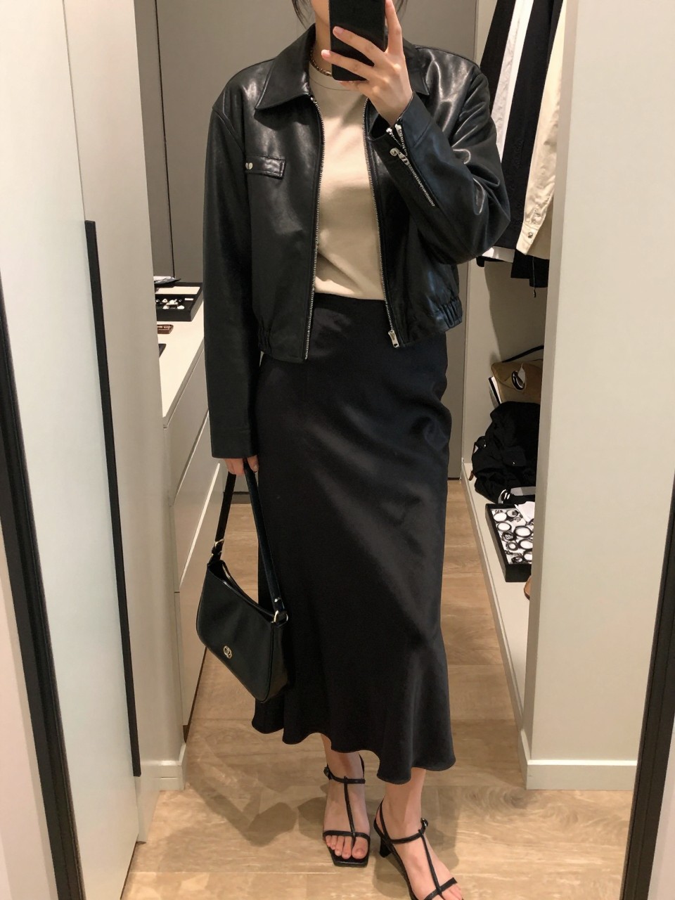 mirror selfie of a woman wearing the exact outfit described above, clearly showing a cropped black fitted leather jacket, soft beige fitted tee, black satin midi slip skirt, black strappy heeled sandals, and a small smooth black leather shoulder bag, holding a phone in front of a mirror, cropped at NECK so the face does not show, with a relaxed casual selfie pose, in a walk-in closet mirror, soft indoor lighting, including an open closet with clothes visible and a jewelry tray on the vanity, latest iPhone photo quality.